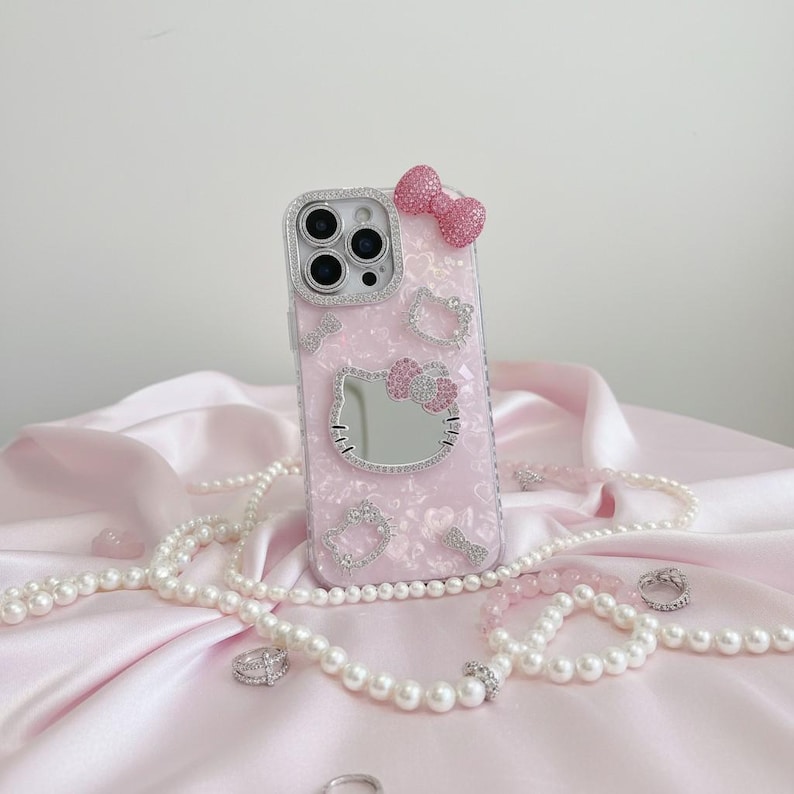 May include: A pink phone case featuring a Hello Kitty design, adorned with rhinestones and a bow. The case includes a mirror and is surrounded by pearl necklaces and rings, displayed on a pink satin fabric.