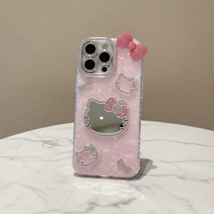 May include: Pink phone case with a Hello Kitty design. The case has a mirror, a bow, and sparkling embellishments. The camera lens is surrounded by a ring of rhinestones. The case has a clear, glittery background and a bow on the top.