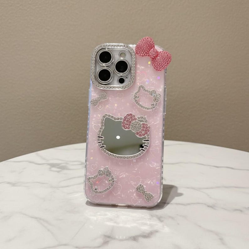 May include: Pink phone case with a Hello Kitty design. The case has a mirror, a bow, and sparkling embellishments. The camera lens is surrounded by a ring of rhinestones. The case has a clear, glittery background and a bow on the top.