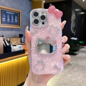 May include: Pink phone case with a Hello Kitty design. The case features a sparkly bow, rhinestones, and a mirror. The case has a pearlescent finish and a silver camera ring.