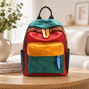 May include: A colorful leather backpack with a patchwork design. The backpack features a teal top, red center, yellow pocket, and green bottom. It has black side pockets, a black handle, and two-tone zipper pulls.