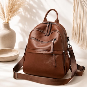 May include: A brown leather backpack with multiple zippered compartments and a top handle. It features adjustable shoulder straps and a crossbody strap. The bag has a textured surface and a classic design, suitable for everyday use.