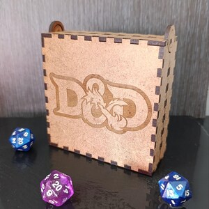 Puede incluir: Una caja de madera clara con el logotipo de D&D grabado, con tres dados de múltiples caras azules y morados. La caja tiene una tapa con bisagras. Los dados están esparcidos.