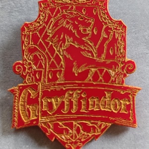 Puede incluir: Un escudo de Gryffindor rojo y dorado. El escudo presenta un león, la palabra "Gryffindor" y detalles ornamentados. El diseño es dorado sobre un fondo rojo. El escudo tiene forma de escudo.
