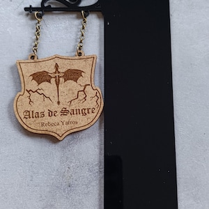 Puede incluir: Un letrero de madera con el texto "Alas de Sangre" y un diseño de dragón, colgado de un soporte de metal negro. El letrero está sujeto con pequeñas cadenas. El letrero también incluye el nombre "Rebecca Yafros".