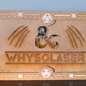 Puede incluir: Una pantalla de juego de mesa de madera con diseños cortados con láser. El panel central presenta el texto "WHYSOLASER" y el logotipo de Dungeons & Dragons. Los paneles laterales tienen ilustraciones de dragones. La pantalla es de madera marrón claro.