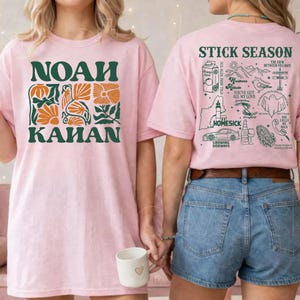 Może przedstawiać: Dwie r&oacute;żowe koszulki. Jedna ma napis "NOAH KAHAN" w kolorze zielonym z pomarańczowymi kwiatowymi akcentami. Druga ma napis "STICK SEASON" i inne ilustracje. Koszulki noszą dwie osoby.