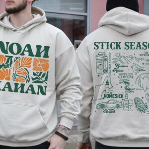 Może przedstawiać: Kremowa bluza z kapturem z zielono-pomarańczową grafiką kwiatową i napisem "NOAH KAHAN" z przodu. Tył przedstawia zielone ilustracje i tekst, w tym "STICK SEASON" i "HOMESICK".