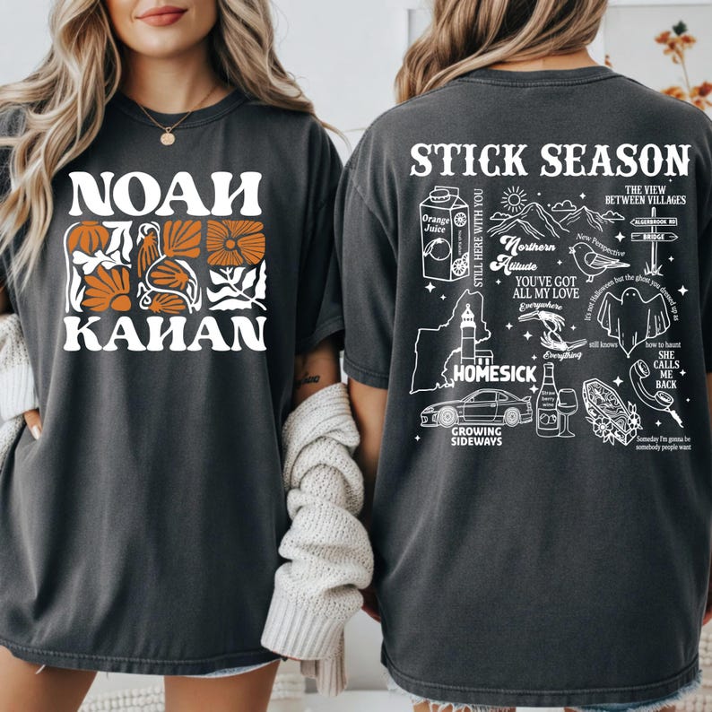 Może przedstawiać: Ciemnoszara koszulka z napisem "NOAH KAHAN" w kolorze białym i pomarańczowym, wraz z ilustracjami kwiat&oacute;w i ptak&oacute;w. Tył koszulki zawiera napis "STICK SEASON" oraz r&oacute;żne elementy graficzne, w tym mapę i samoch&oacute;d.