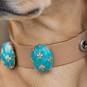 May include: Tan leather dog collar with two turquoise and gold decorative accents. The turquoise accents are oval-shaped and feature a gold lattice design with pink gemstone embellishments. The collar has a silver grommet.