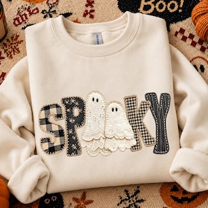May include: A cream-coloured crewneck sweatshirt featuring the word SPOOKY across the chest. The letters are appliqued with various fabrics including black and white buffalo plaid, black floral print, and two white lace ghost figures.