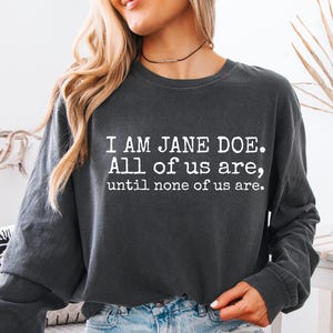 May include: A charcoal gray long-sleeve crewneck sweatshirt featuring white text that reads: I AM JANE DOE. All of us are, until none of us are. The garment has a relaxed fit with ribbed cuffs and a ribbed neckline, made of a soft fabric.