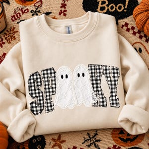 May include: A cream-coloured crewneck sweatshirt featuring the word SPOOKY appliqued on the front. The letters S, P, K, and Y are made of black and white buffalo plaid fabric, while the two O's are replaced by white lace ghost figures.