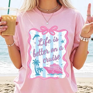 May include: Pink t-shirt with the phrase "Life is better on a cruise" in a decorative frame. The graphic includes a cruise ship, palm trees, a dolphin, and a star. The shirt is a light pink color.