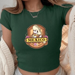 May include: Dark green crop top featuring a cartoon character wearing a sombrero, with the word "MEXICO" in a decorative banner. The design includes flowers and the text "FOOD, FRIENDS, FIESTA".