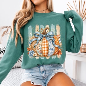 May include: A teal long-sleeve sweatshirt featuring a central graphic of a pumpkin decorated with various patterns including leopard print, orange gingham, and blue floral designs, tied with a large blue ribbon and surrounded by autumn leaves.