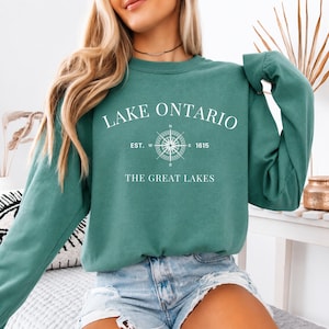 May include: A teal long-sleeve crewneck sweatshirt featuring white text that reads LAKE ONTARIO, EST. 1615, and THE GREAT LAKES, centred around a white compass graphic. The sweatshirt has a ribbed neckline, cuffs, and hem, with a solid colour finish.