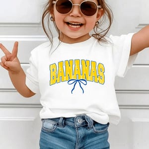 May include: White t-shirt with the word "BANANAS" in yellow and blue lettering, and a blue bow graphic. The shirt is paired with blue jeans. The person is wearing sunglasses and making a peace sign.