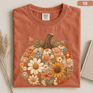May include: A terracotta-coloured crew neck t-shirt featuring a large, central graphic of a pumpkin filled with embroidered-style floral patterns in shades of orange, yellow, white, and blue. The shirt has a Comfort Colors brand label at the neck.