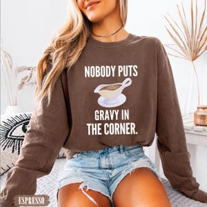 May include: A brown long-sleeve crewneck sweatshirt featuring a white graphic of a gravy boat on a saucer with the text NOBODY PUTS GRAVY IN THE CORNER printed in white capital letters above and below the illustration of the gravy boat.