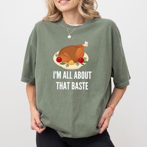 May include: An olive green short-sleeve t-shirt featuring a graphic of a roasted turkey on a platter with red tomatoes and the white text "I'M ALL ABOUT THAT BASTE" printed underneath the illustration in a clean, sans-serif font.