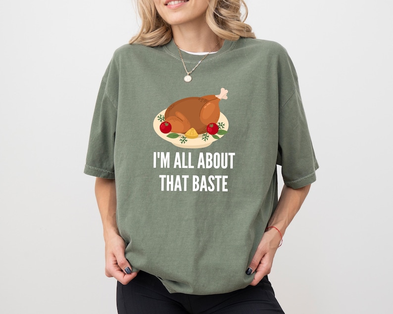 May include: An olive green short-sleeve t-shirt featuring a graphic of a roasted turkey on a platter with red tomatoes and the white text "I'M ALL ABOUT THAT BASTE" printed underneath the illustration in a clean, sans-serif font.