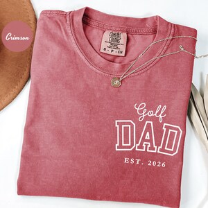 May include: Crimson-coloured t-shirt with the words "Golf Dad" in white block letters and "EST. 2026" below. A gold necklace with a pendant rests on the shirt. The shirt has a crew neck and a small tag.