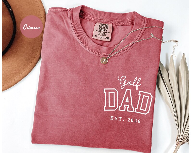 May include: Crimson-coloured t-shirt with the words "Golf Dad" in white block letters and "EST. 2026" below. A gold necklace with a pendant rests on the shirt. The shirt has a crew neck and a small tag.