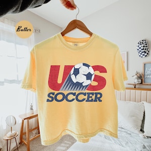 Peut inclure: T-shirt jaune clair avec le texte "US SOCCER" en rouge et bleu. Le motif repr&eacute;sente un ballon de football avec une tra&icirc;n&eacute;e bleue et blanche. Le t-shirt est suspendu &agrave; un cintre en bois.