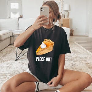 May include: A black short-sleeved t-shirt featuring a graphic of a slice of pumpkin pie topped with a dollop of white whipped cream. Below the pie graphic, the text PIECE OUT is printed in white capital letters in a simple sans-serif font.