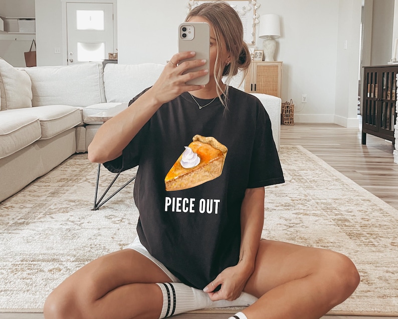 May include: A black short-sleeved t-shirt featuring a graphic of a slice of pumpkin pie topped with a dollop of white whipped cream. Below the pie graphic, the text PIECE OUT is printed in white capital letters in a simple sans-serif font.