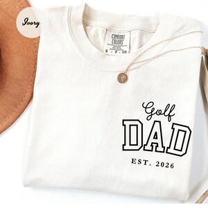 May include: Ivory t-shirt featuring the text "Golf DAD EST. 2026" in black block letters. The shirt has a crew neck and short sleeves. A gold necklace with a pendant is draped across the shirt.
