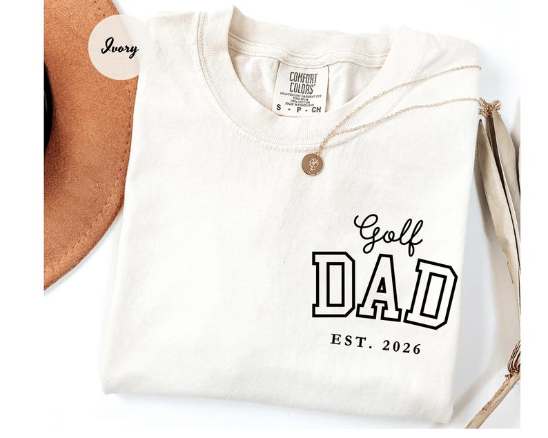 May include: Ivory t-shirt featuring the text "Golf DAD EST. 2026" in black block letters. The shirt has a crew neck and short sleeves. A gold necklace with a pendant is draped across the shirt.