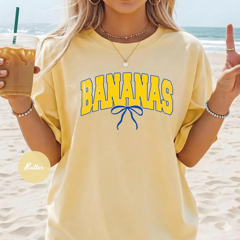 May include: Yellow t-shirt with the word "BANANAS" in blue and yellow lettering. A blue bow is beneath the word. The shirt is a light yellow color.