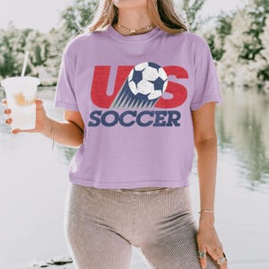 Peut inclure: Un crop top lilas avec le texte "US SOCCER" en rouge et bleu marine. Le motif pr&eacute;sente un ballon de football avec une tra&icirc;n&eacute;e de lignes bleues. Le t-shirt est en mati&egrave;re souple.