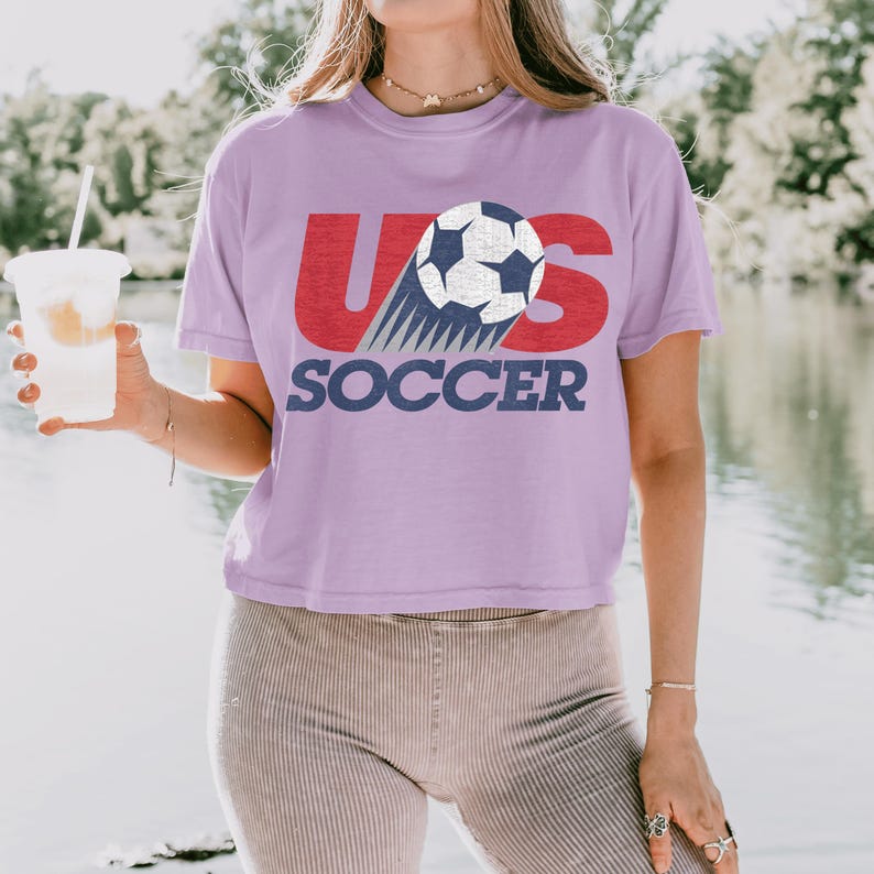 Peut inclure: Un crop top lilas avec le texte "US SOCCER" en rouge et bleu marine. Le motif pr&eacute;sente un ballon de football avec une tra&icirc;n&eacute;e de lignes bleues. Le t-shirt est en mati&egrave;re souple.