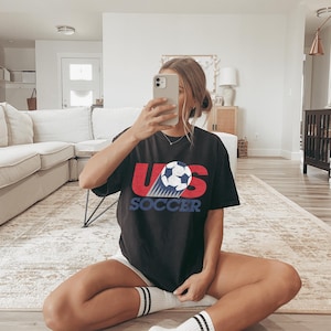 May include: Black t-shirt with the text "US SOCCER" in red and blue, featuring a soccer ball graphic. The shirt is a casual style, suitable for sports fans.