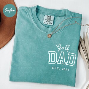 May include: Seafoam green t-shirt with the text "Golf DAD EST. 2026" in white block letters. A gold necklace with a pendant rests on the shirt. The shirt is folded and has a Comfort Colors tag.
