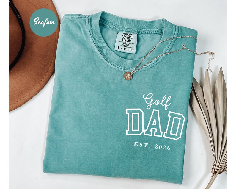 May include: Seafoam green t-shirt with the text "Golf DAD EST. 2026" in white block letters. A gold necklace with a pendant rests on the shirt. The shirt is folded and has a Comfort Colors tag.