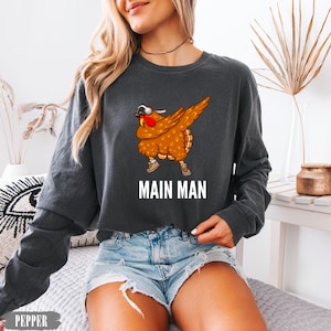 May include: A charcoal gray long-sleeve crewneck sweatshirt featuring a graphic of a brown turkey wearing sunglasses and sneakers while performing a dab pose. Below the turkey, the text MAIN MAN is printed in bold white capital letters.