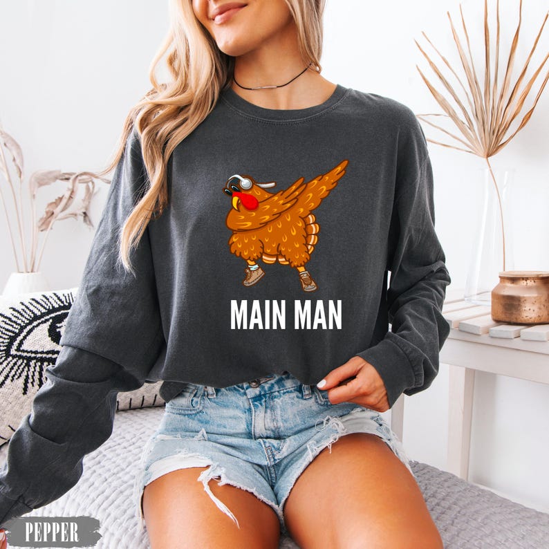 May include: A charcoal gray long-sleeve crewneck sweatshirt featuring a graphic of a brown turkey wearing sunglasses and sneakers while performing a dab pose. Below the turkey, the text MAIN MAN is printed in bold white capital letters.