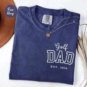 May include: A navy blue t-shirt with the words "Golf DAD EST. 2026" printed in white. The shirt has a crew neck and a small label on the collar. A gold necklace with a pendant is draped across the shirt.