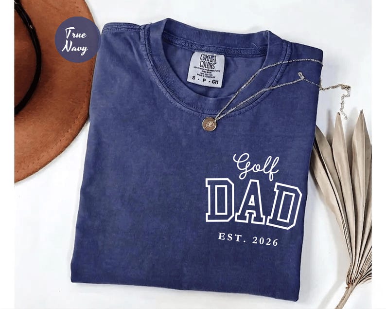 May include: A navy blue t-shirt with the words "Golf DAD EST. 2026" printed in white. The shirt has a crew neck and a small label on the collar. A gold necklace with a pendant is draped across the shirt.