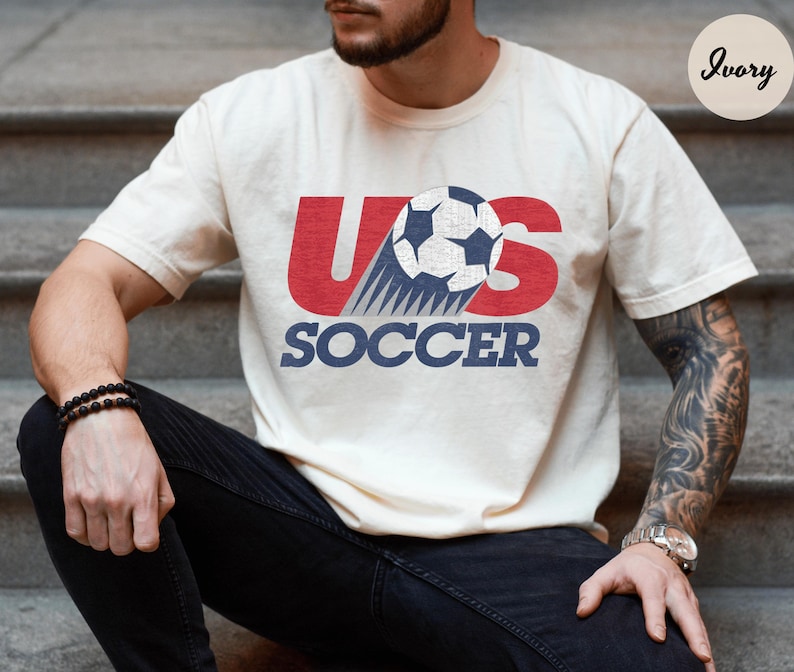 May include: Ivory-colored t-shirt with the text "US SOCCER" in red and blue. The design features a soccer ball graphic. The shirt is short-sleeved and made of a soft material.