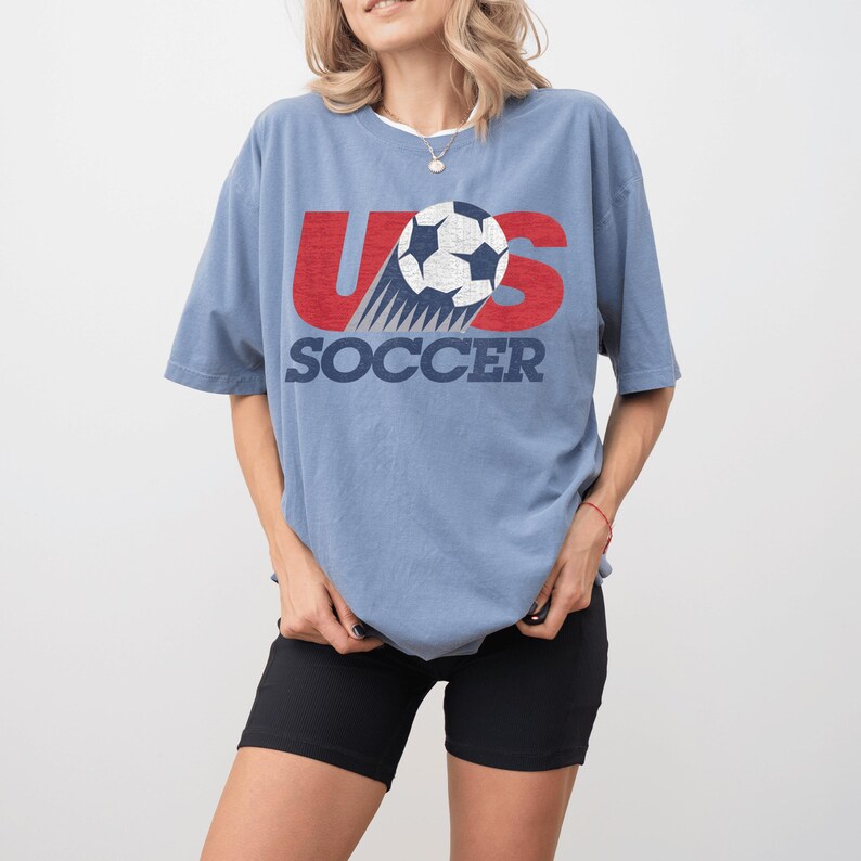 May include: A light blue t-shirt with the text "US SOCCER" in red and navy blue. The design features a soccer ball with motion lines. The shirt is a casual style.
