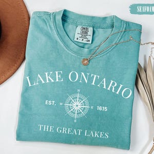 May include: A seafoam green short-sleeve t-shirt featuring white text that reads LAKE ONTARIO, EST. 1615, and THE GREAT LAKES, along with a white compass graphic. The shirt has a Comfort Colors brand label at the neck and a size small tag.