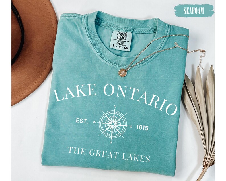 May include: A seafoam green short-sleeve t-shirt featuring white text that reads LAKE ONTARIO, EST. 1615, and THE GREAT LAKES, along with a white compass graphic. The shirt has a Comfort Colors brand label at the neck and a size small tag.