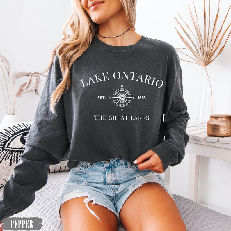 May include: A charcoal grey long-sleeve sweatshirt featuring white text that reads LAKE ONTARIO, a compass graphic, EST. 1615, and THE GREAT LAKES. The garment has a crew neckline and a relaxed fit, with the colour name PEPPER printed in the corner.