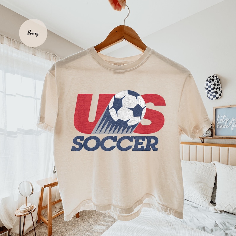 Peut inclure: Un t-shirt cr&egrave;me avec le texte "US SOCCER" en rouge et bleu. Un graphisme de ballon de football est incorpor&eacute; dans le design. Le t-shirt est suspendu &agrave; un cintre en bois.