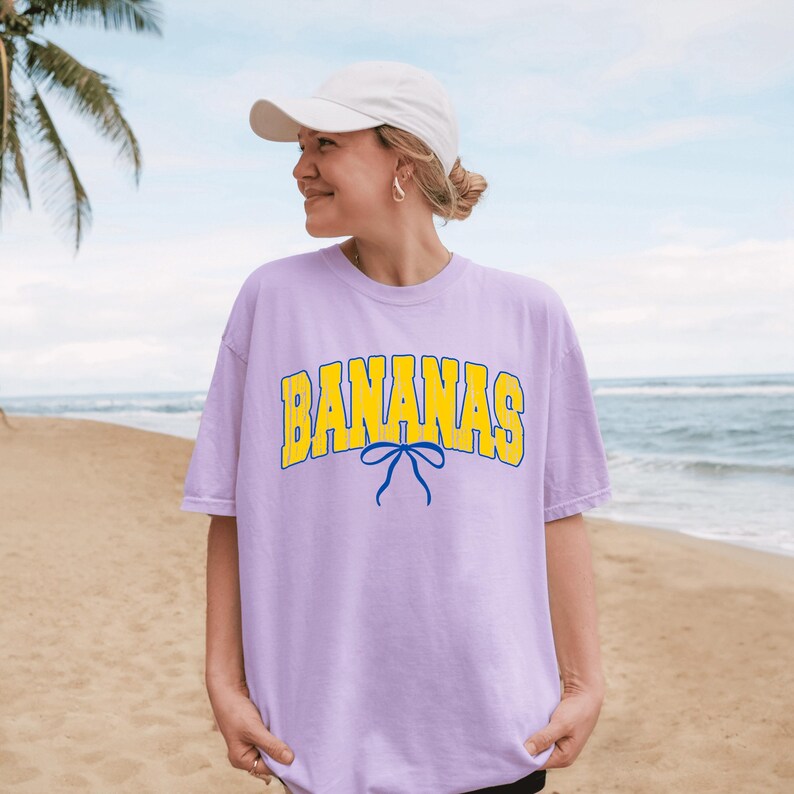 May include: A light purple t-shirt with the word "BANANAS" in yellow block letters with a blue bow graphic. The shirt is worn with a white baseball cap on a beach with palm trees and the ocean in the background.